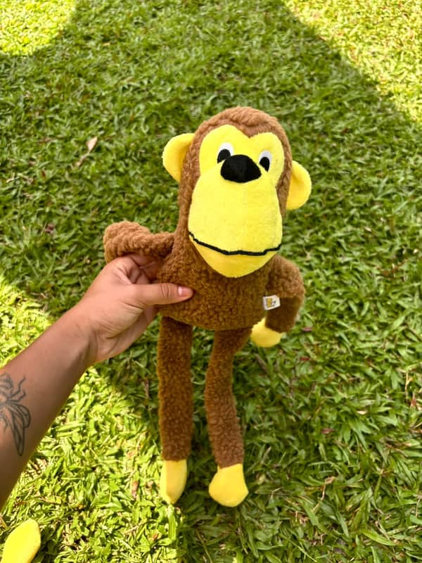 Macaco de pelúcia marrom com rosto, mãos e pés amarelos, braços e pernas compridos
